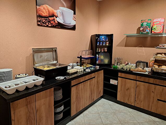 Frühstückbuffet in Holzoptik mit schwarzer Arbeitsplatte, weißer Besteckkorb, verschiedene Teller, Chafing Dish mit Eier, Speck und Würstchen, Eierkocher, Osttiroler Eier, Etager mit verschiedenen Wurst- und Käsesorten, Bresso, Brunch, schwarzer Kühlschrank mit verschiedenen Joghurt zum Beispiel Cremix, Tirolmilch Naturjoghurt, Fruchtjoghurt, anschließend Fleischaufstriche, veganen Aufstrich, Brot, Toaster, Etager mit verschiedenen Brot und Gebäcksorten, darüber Kellogscerealien, außerdem ein Frühstücksbild